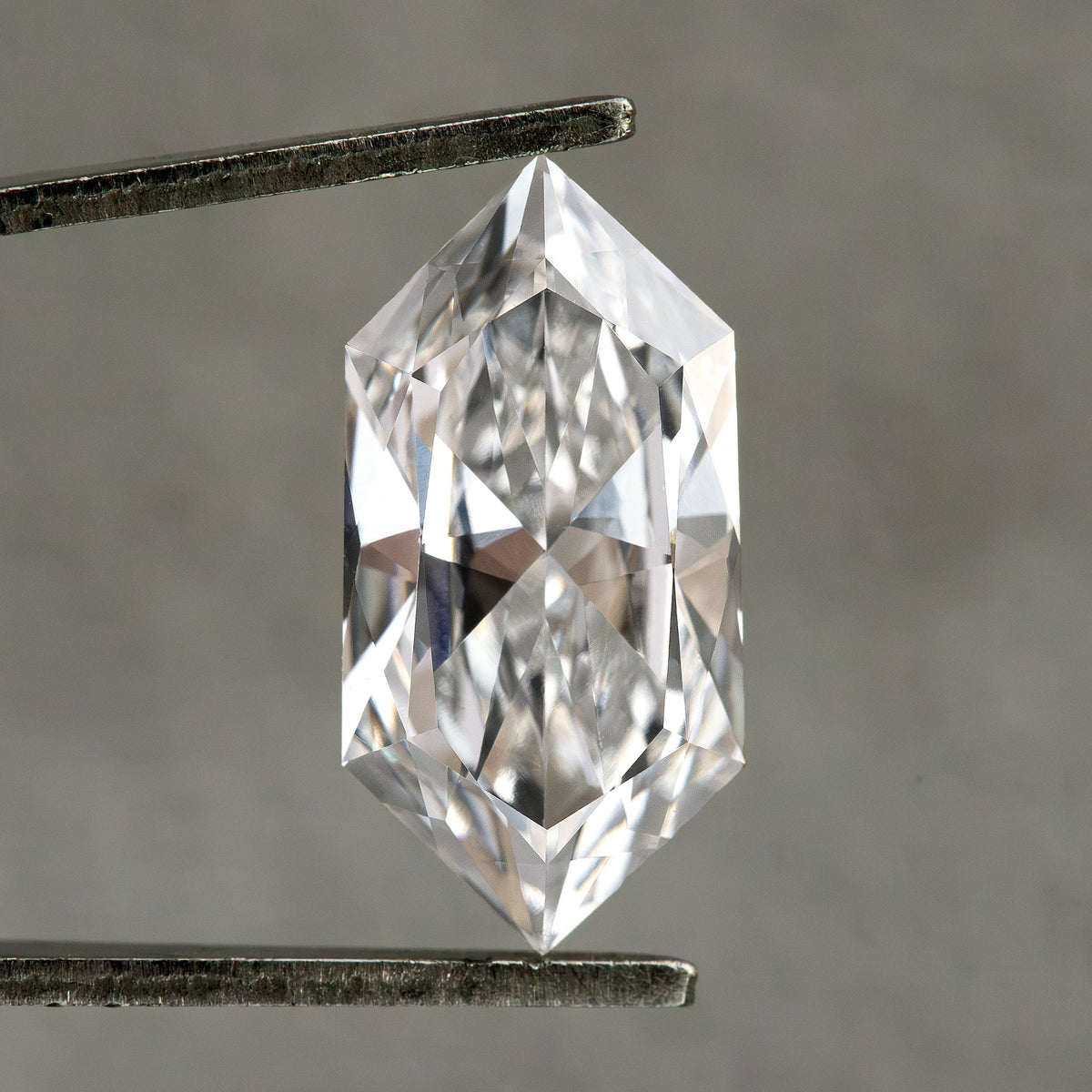 Studio face-up view of an elongated hexagonal diamond held vertically in tweezers on neutral gray background.
