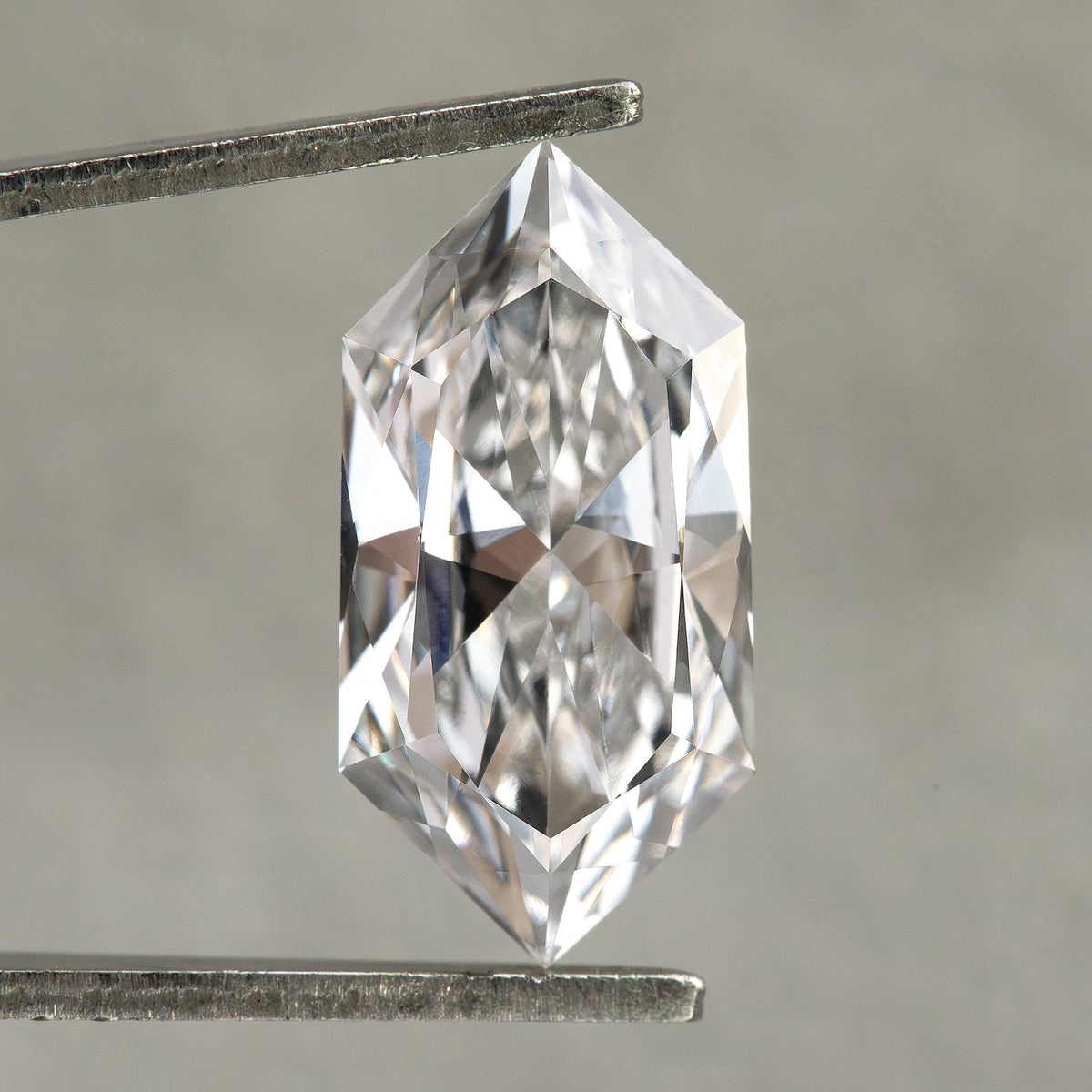 Colorless elongated hexagon cut diamond held in tweezers under crisp illumination against gray background.