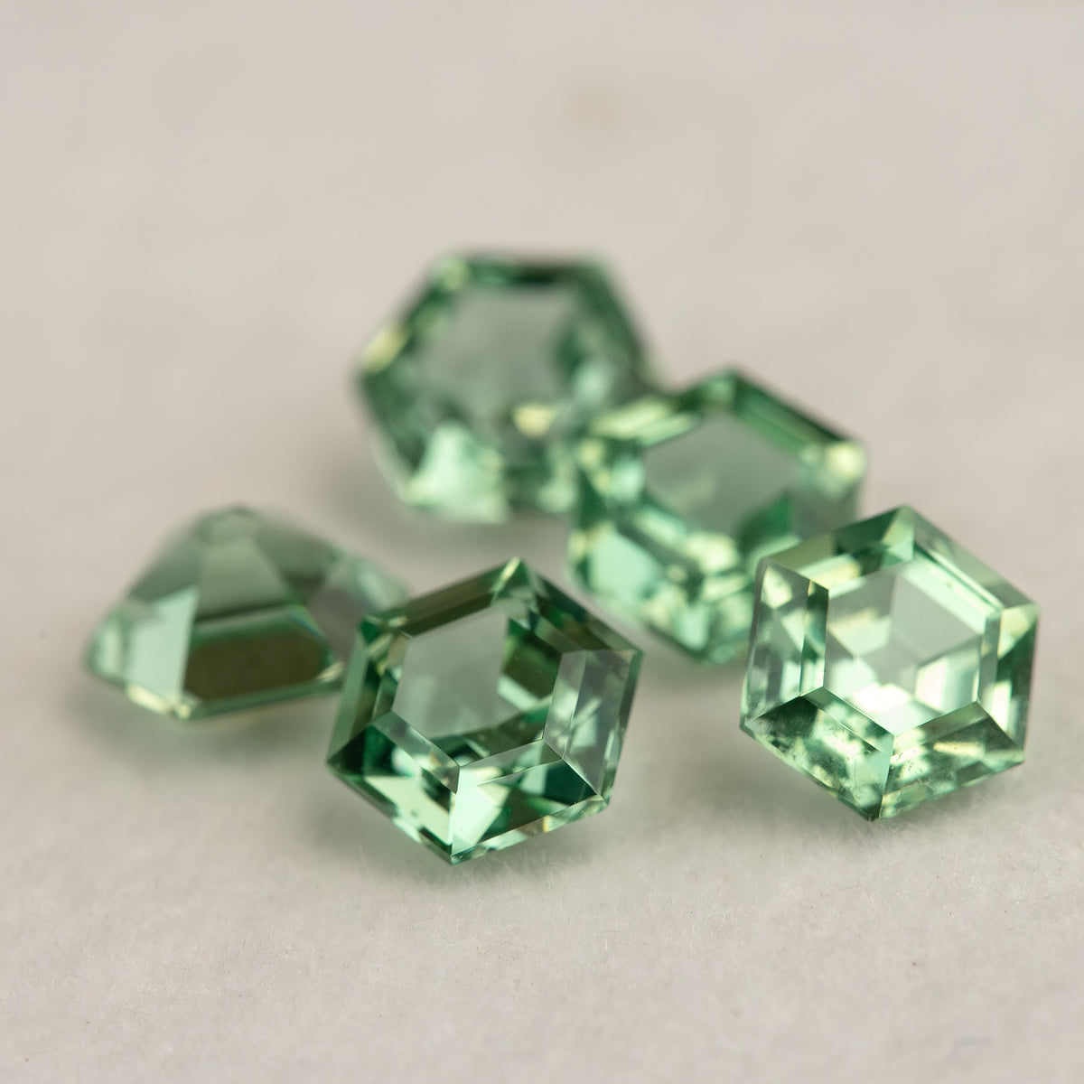 Five mint green hexagonal lab-grown sapphires grouped on a pale surface, with one stone slightly out of focus behind the others.
