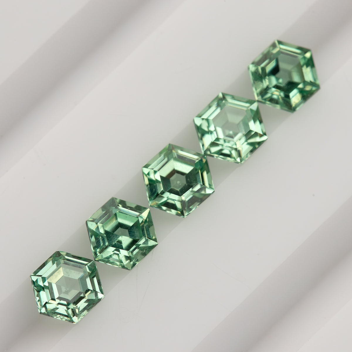 Five mint green hexagonal lab-grown sapphires in a diagonal row on a white grooved tray.