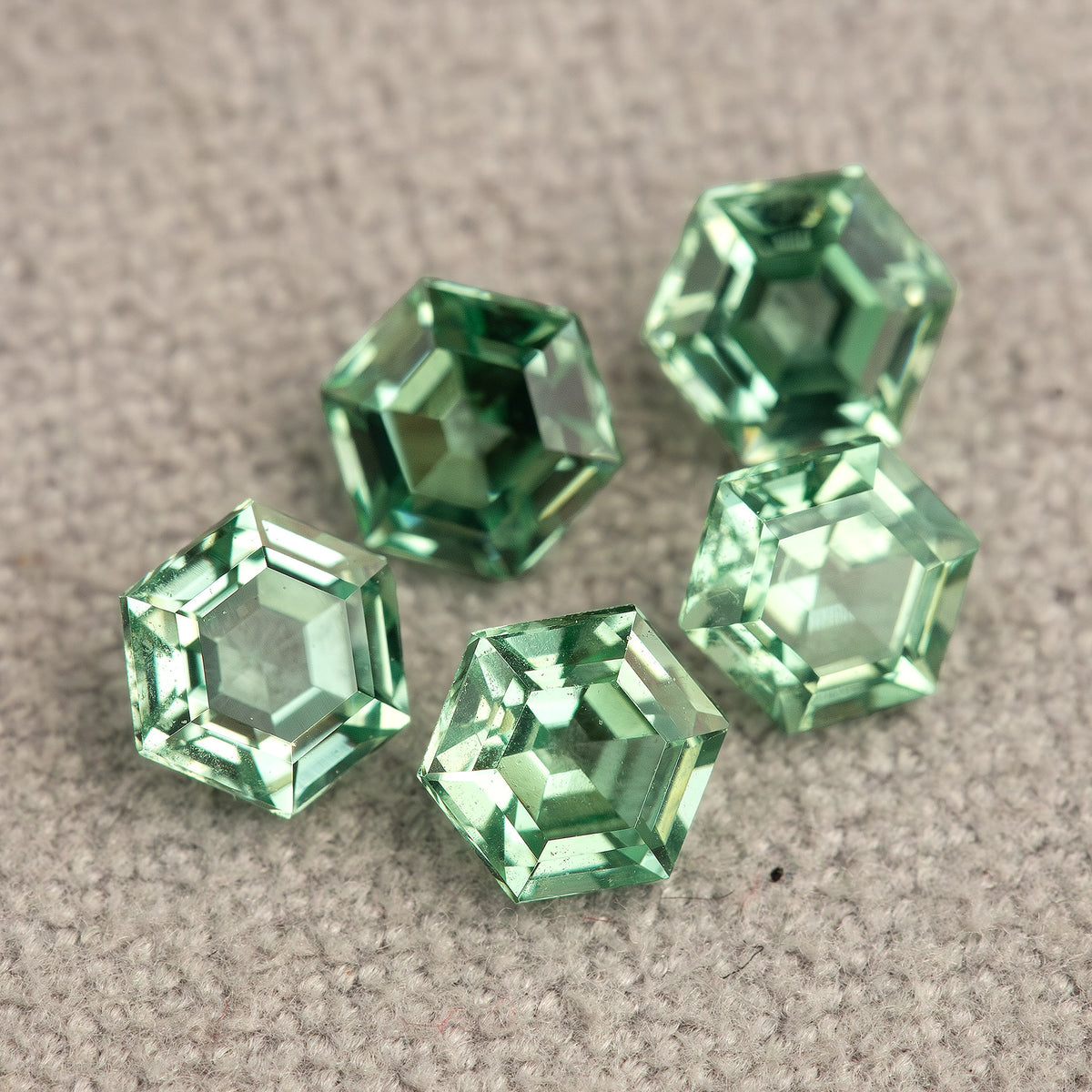 Mint green hexagonal lab-grown sapphires grouped on textured pale grey fabric, with contrasting bright and dark reflections.