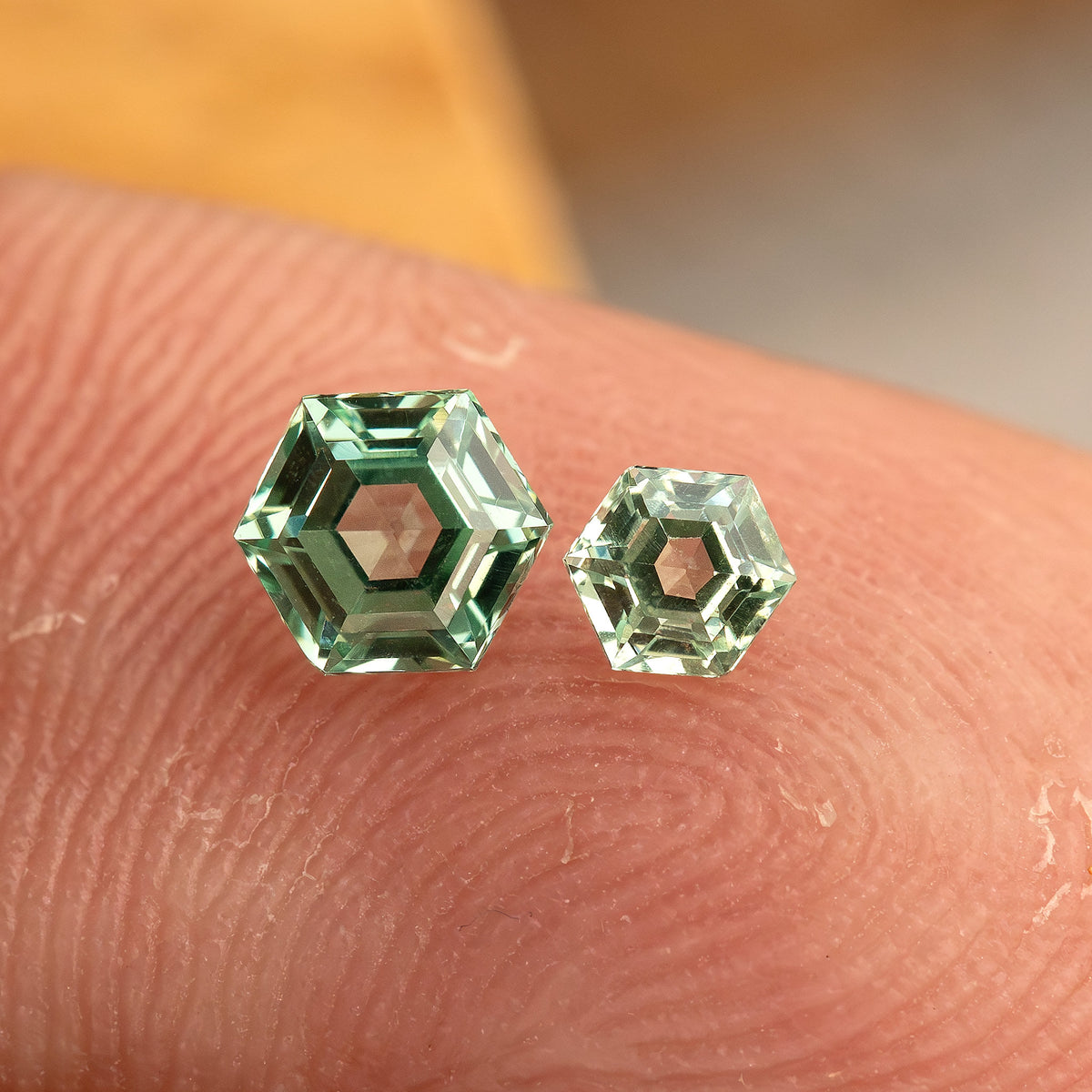 Two mint green hexagonal lab-grown sapphires of different sizes resting face-up on a fingertip for relative scale.