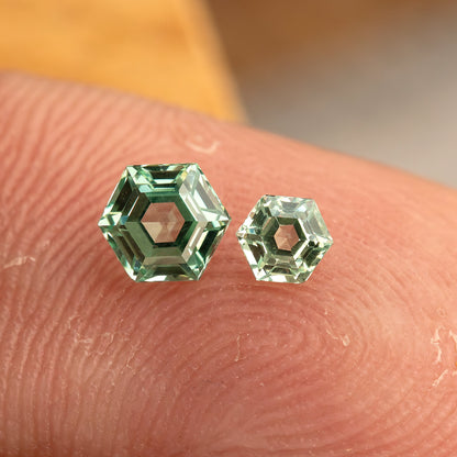 Two mint green hexagonal lab-grown sapphires of different sizes resting face-up on a fingertip for relative scale.