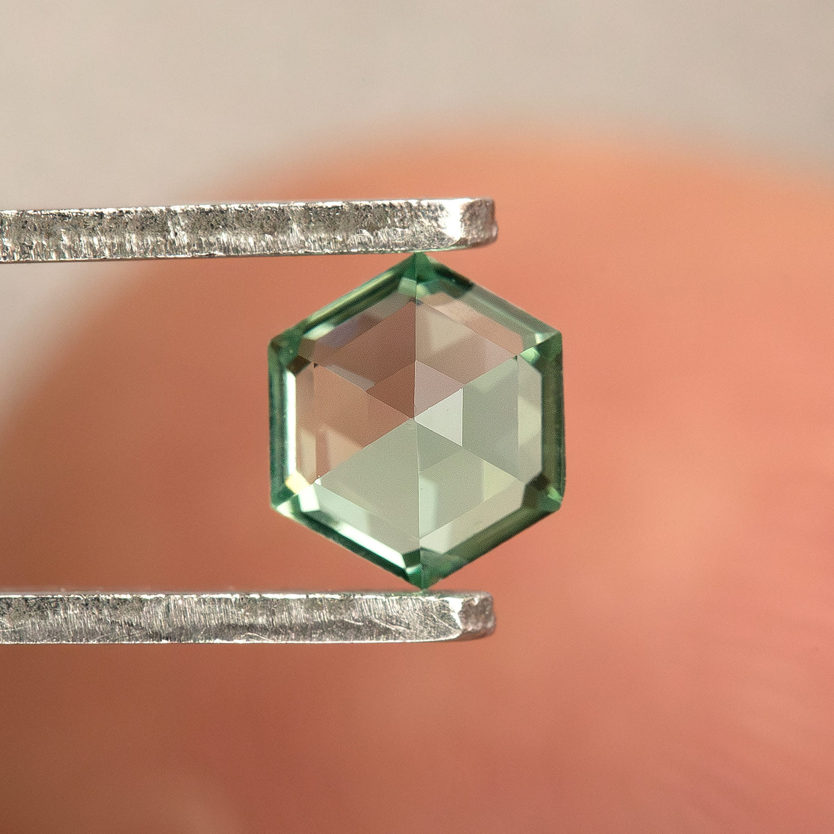Opposite-side view of a mint green hexagonal lab-grown sapphire in tweezers, showing facets converging toward the center.