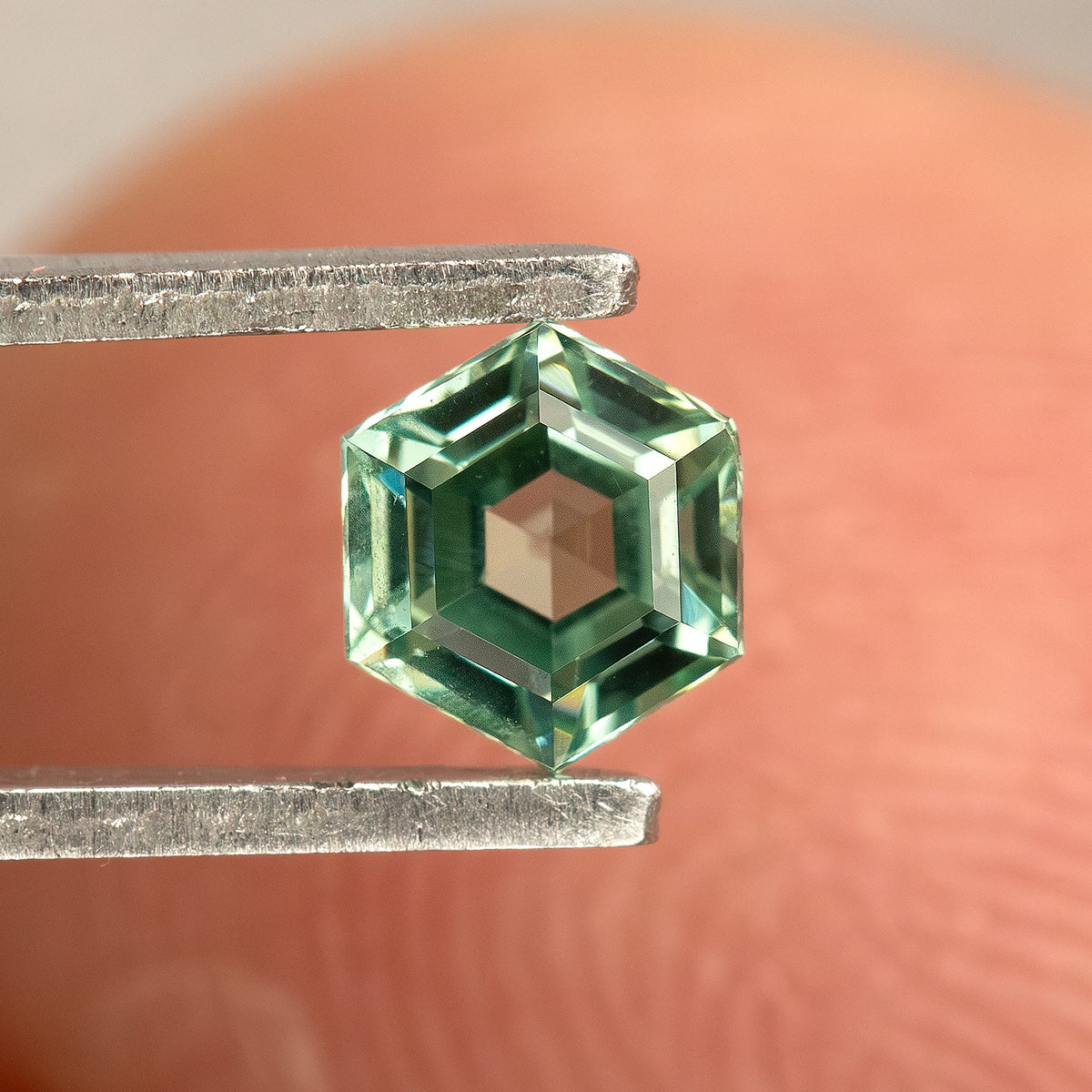 Face-up mint green hexagonal lab-grown sapphire in tweezers, showing its flat central table and surrounding facets.