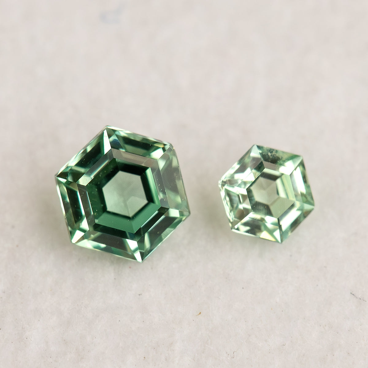 Larger and smaller mint green hexagonal lab-grown sapphires face-up on a white surface, showing flat tables and contrasting green reflections.
