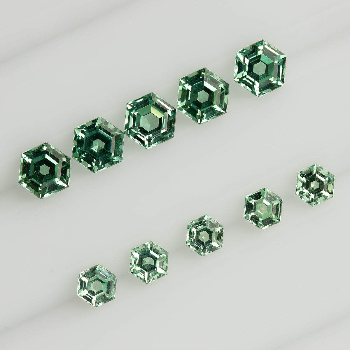 Larger and smaller mint green hexagonal lab-grown sapphires arranged in two diagonal rows on a white tray.