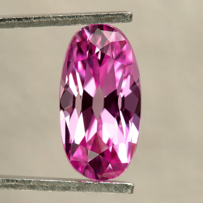 Hot-pink lab-grown Moval Old Mine Cut sapphire held upright face-up in tweezers, showing its elongated rounded outline and alternating bright pink and dark facets.