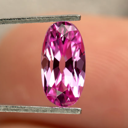 Pink lab-grown moval sapphire in tweezers in front of a fingertip, with a shifted pattern of pale pink and fuchsia reflections.