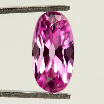 Neutral-background face-up view of the hot-pink moval sapphire in tweezers, showing its rounded ends and broad reflective crown facets.