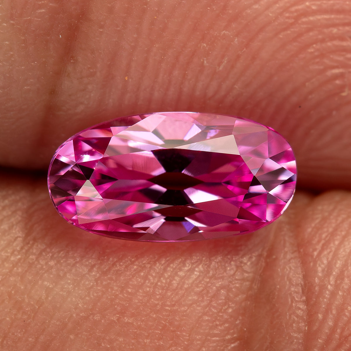 Pink moval-cut lab-grown sapphire resting horizontally between fingertips, with skin texture showing the representative stone scale.