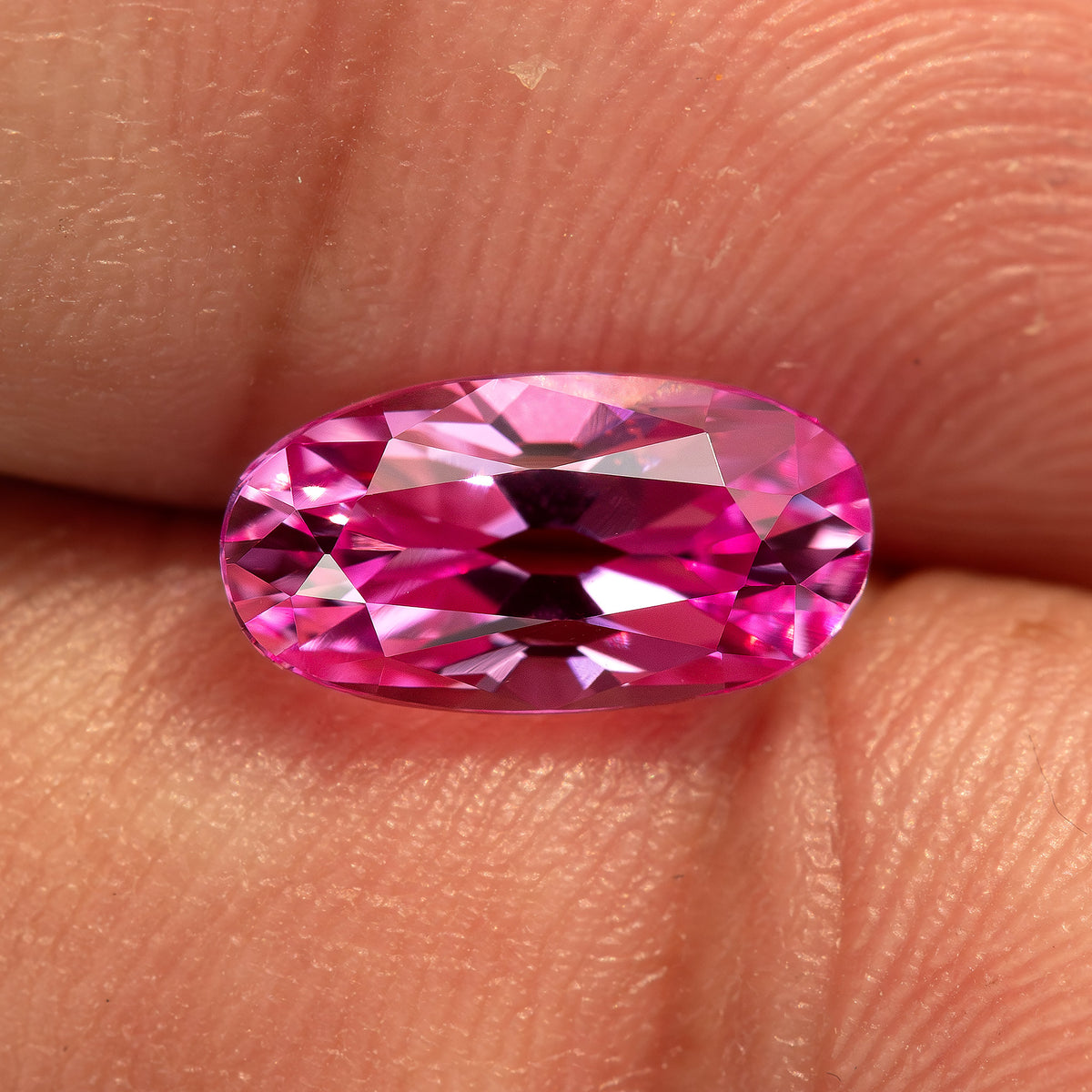 Hot-pink lab-grown sapphire on fingertips at a brighter angle, showing its elongated moval outline and changed facet reflections.