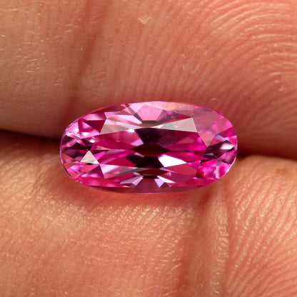 Hot-pink lab-grown sapphire on fingertips at a brighter angle, showing its elongated moval outline and changed facet reflections.