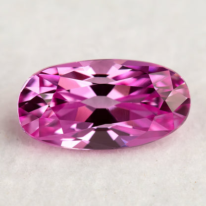 Hot-pink moval sapphire on a pale textured surface, showing bright crown reflections and its elongated rounded outline.