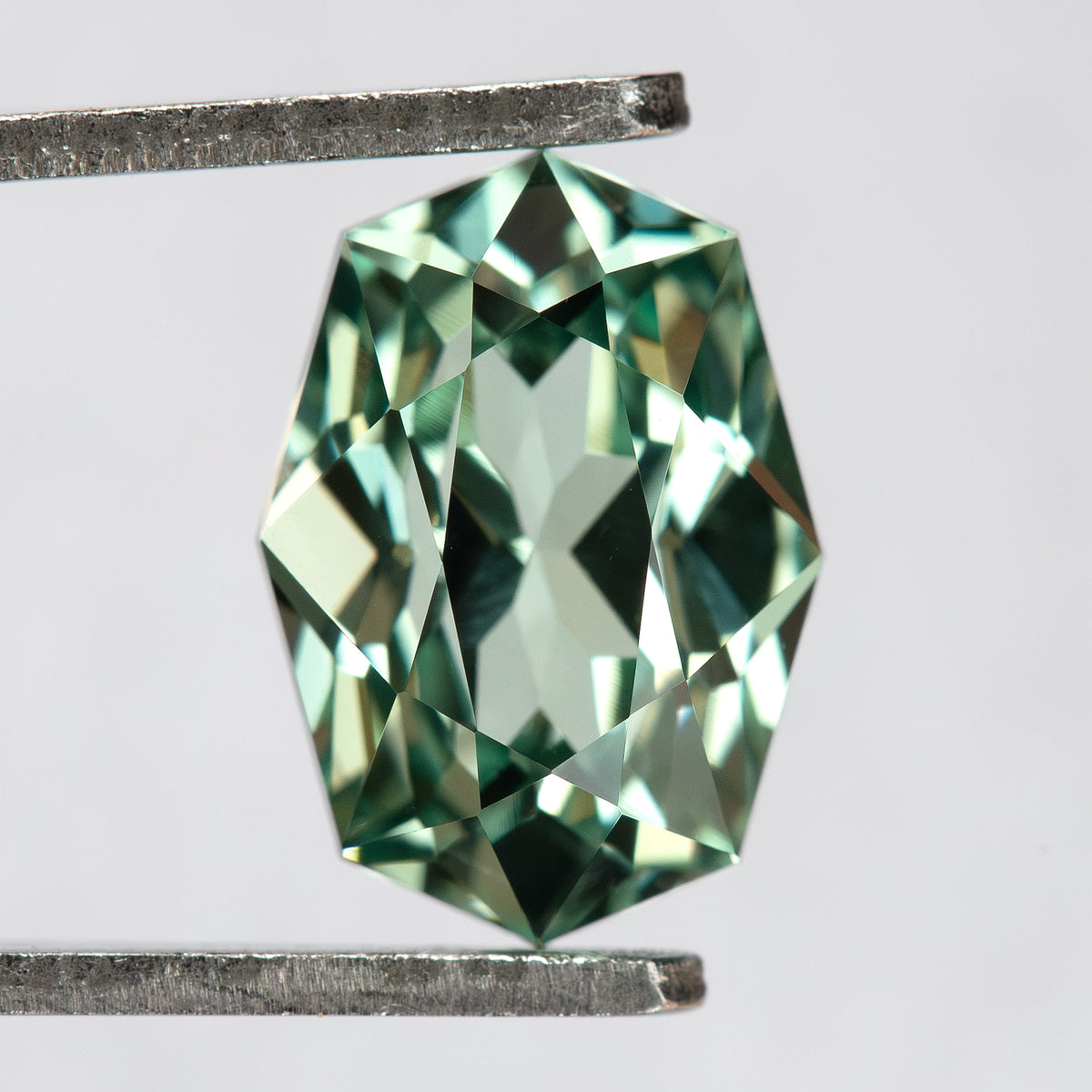 Mint green lab-grown sapphire in tweezers, showing the BLS111 elongated-octagon face-up outline and geometric facet pattern.