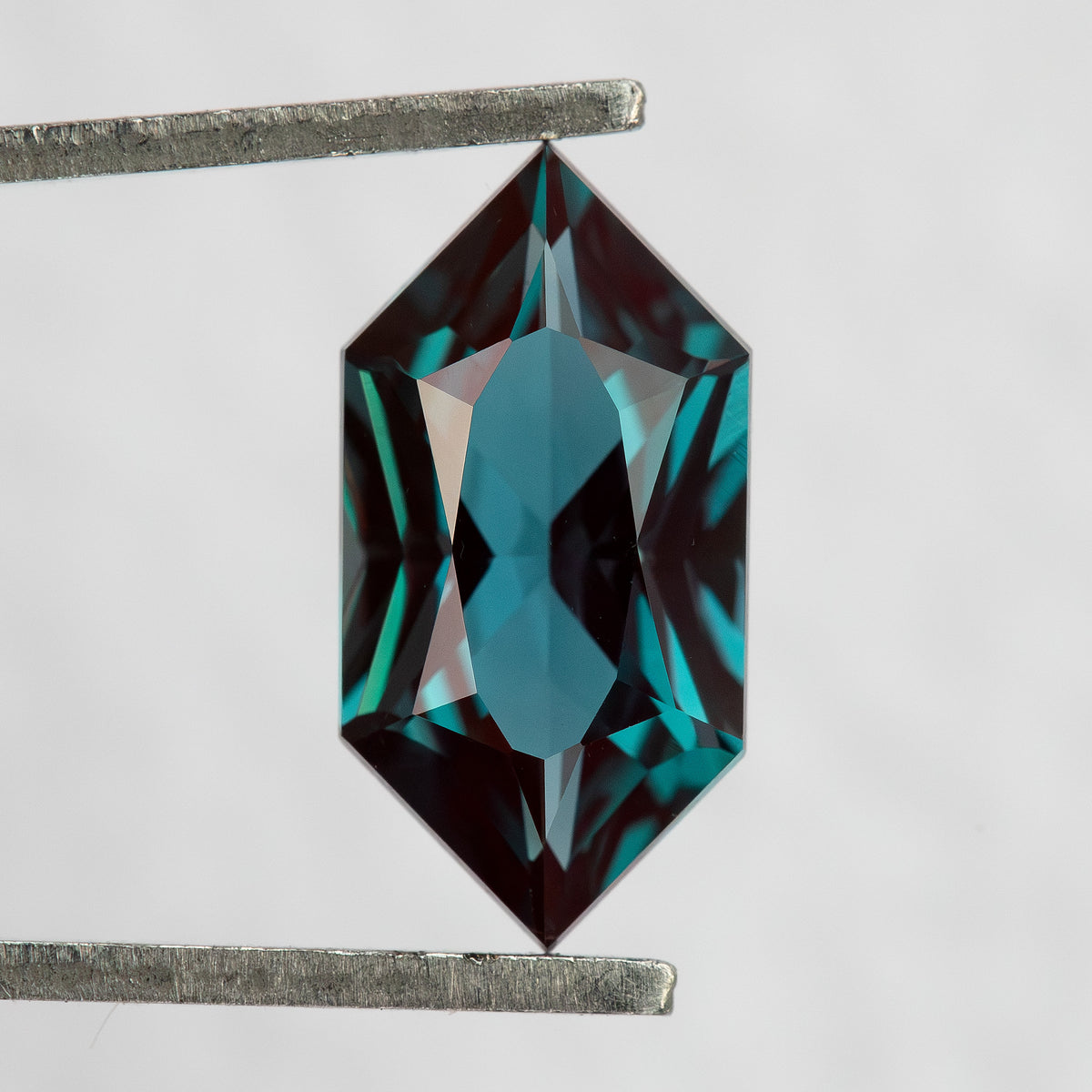 Face-up BLS113 lab-grown alexandrite in tweezers, showing its Dutch Marquise Hexagon outline and greenish blue color.