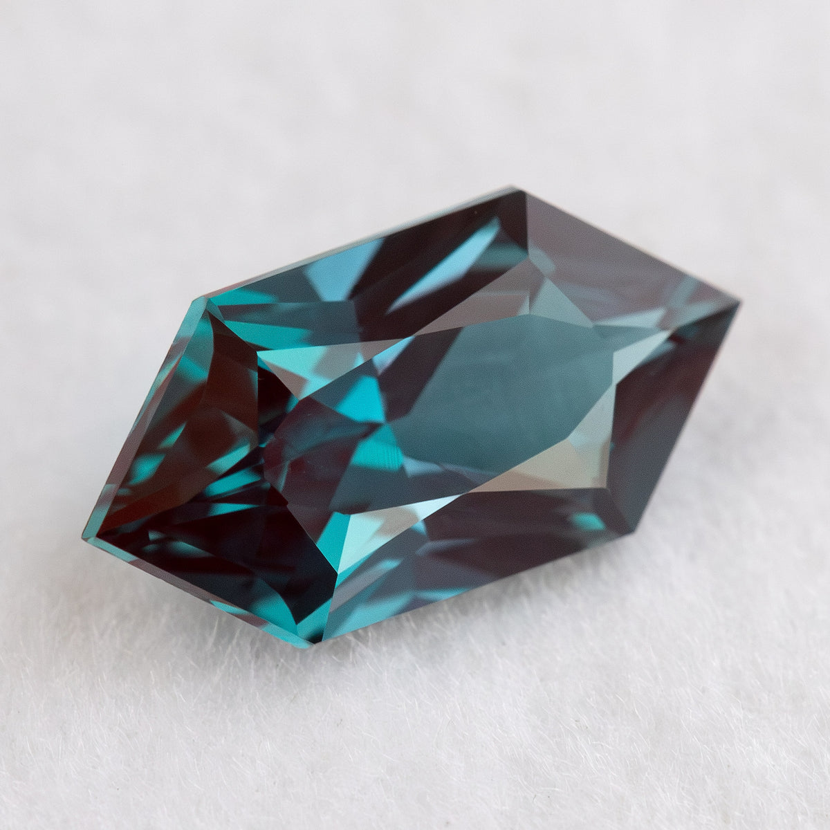 Angled view of BLS113 lab-grown alexandrite on white, showing greenish blue color and red-purple reflections.