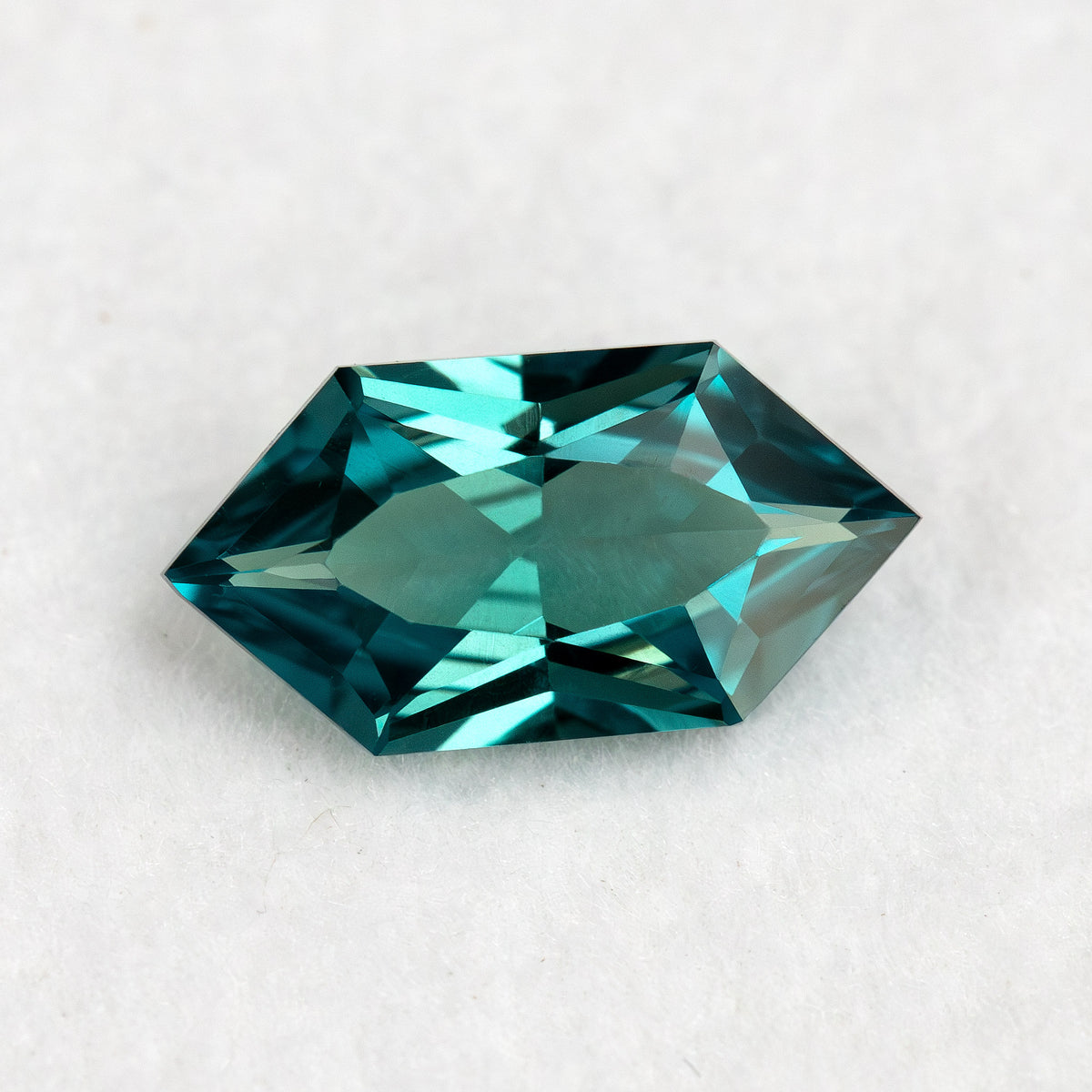 Face-up BLS115 teal green lab-grown sapphire on white, showing its Dutch Marquise Hexagon outline and geometric facets.