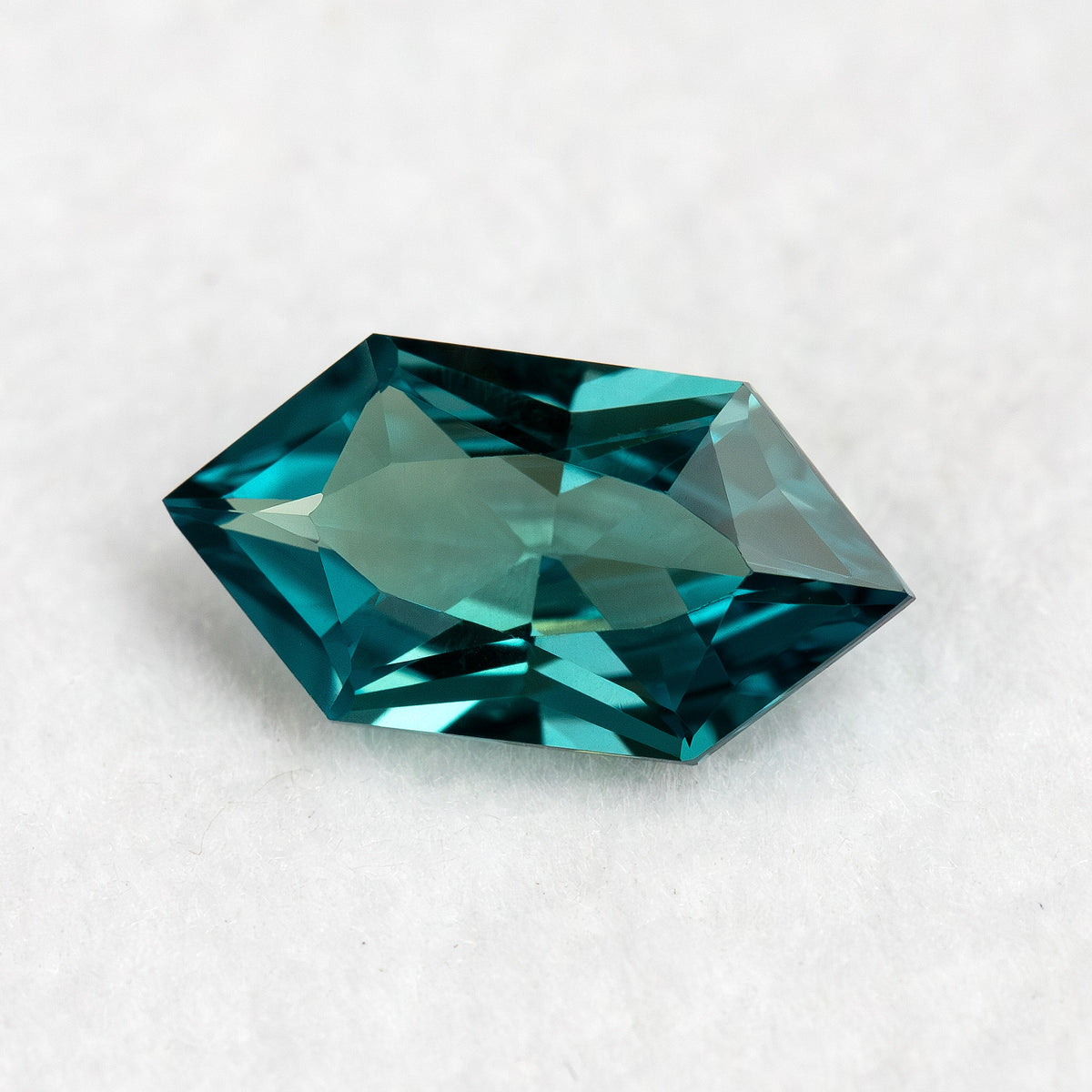 Angled face-up view of BLS115 lab-grown sapphire on white, showing teal green color and an elongated hexagonal outline.
