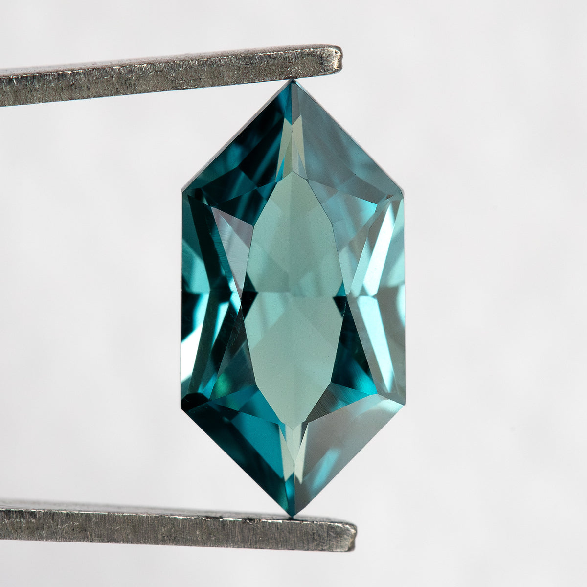 Face-up BLS115 teal green lab-grown sapphire held vertically in tweezers, showing its Dutch Marquise Hexagon outline.