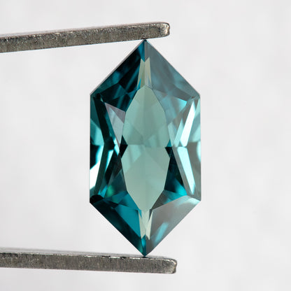 Face-up BLS115 teal green lab-grown sapphire held vertically in tweezers, showing its Dutch Marquise Hexagon outline.
