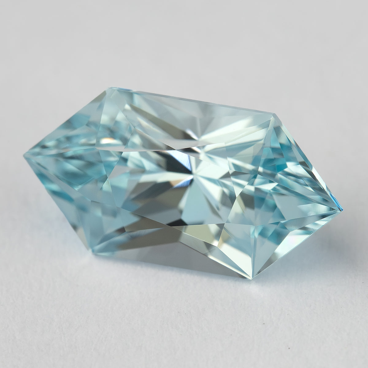 Slightly angled face-up view of an aquamarine-blue lab-grown sapphire with an elongated hexagon outline and geometric facets on a pale background.