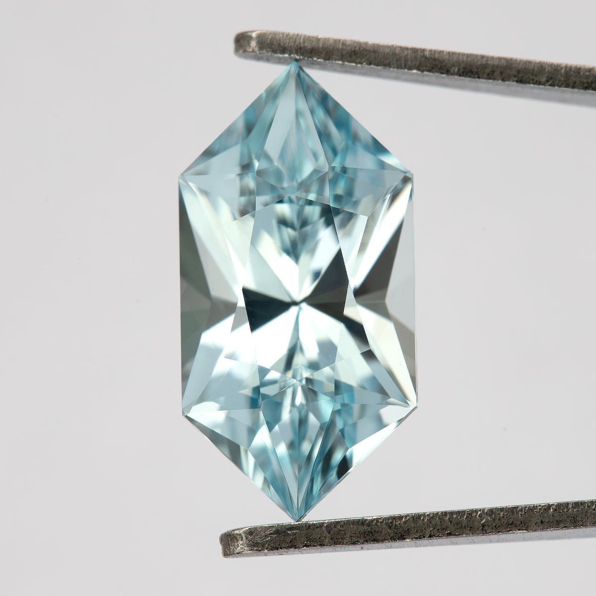 Aquamarine-blue lab-grown sapphire held upright in tweezers, showing its face-up facets and elongated hexagon outline.