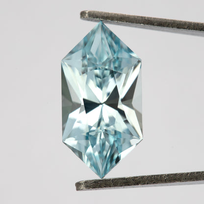Aquamarine-blue lab-grown sapphire held upright in tweezers, showing its face-up facets and elongated hexagon outline.