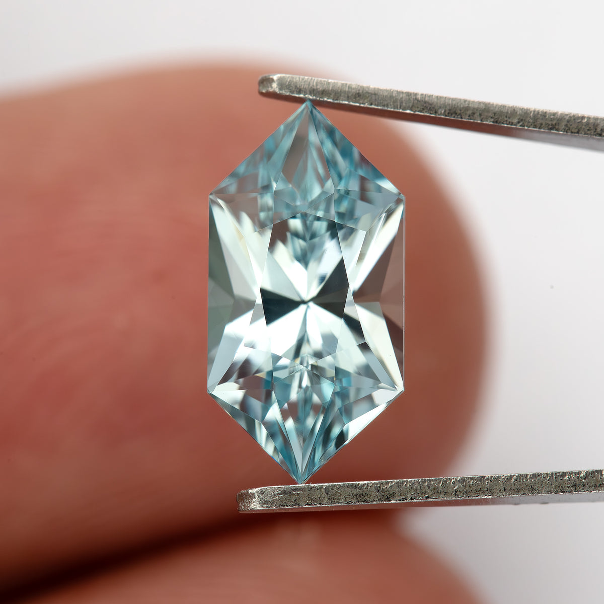Upright face-up view of the lab-grown sapphire in tweezers against a fingertip, showing its pointed ends.