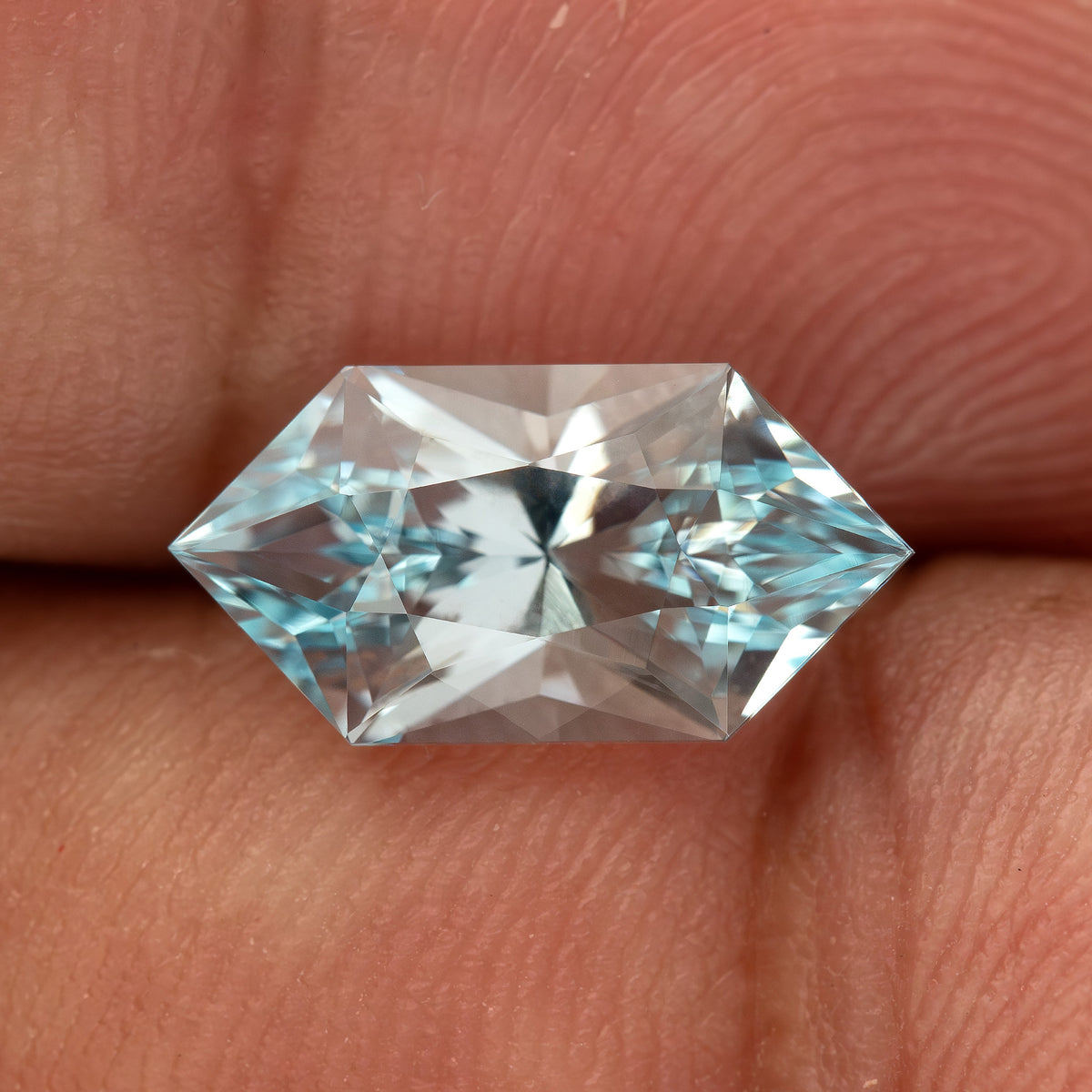 Pale-blue lab-grown sapphire on a fingertip, with bright reflections across its elongated hexagon face.
