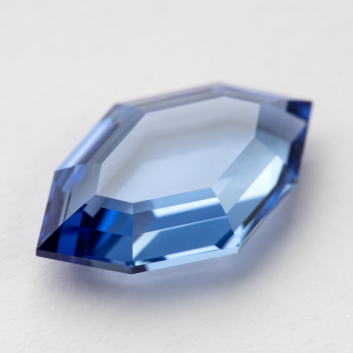 Low tilted view of the blue portrait-cut sapphire on white, showing bright step reflections along the edges.