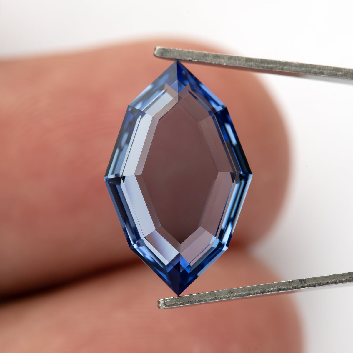 Blue portrait-cut sapphire held face-up vertically in tweezers against a fingertip for scale.