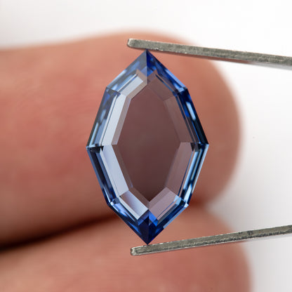 Blue portrait-cut sapphire held face-up vertically in tweezers against a fingertip for scale.