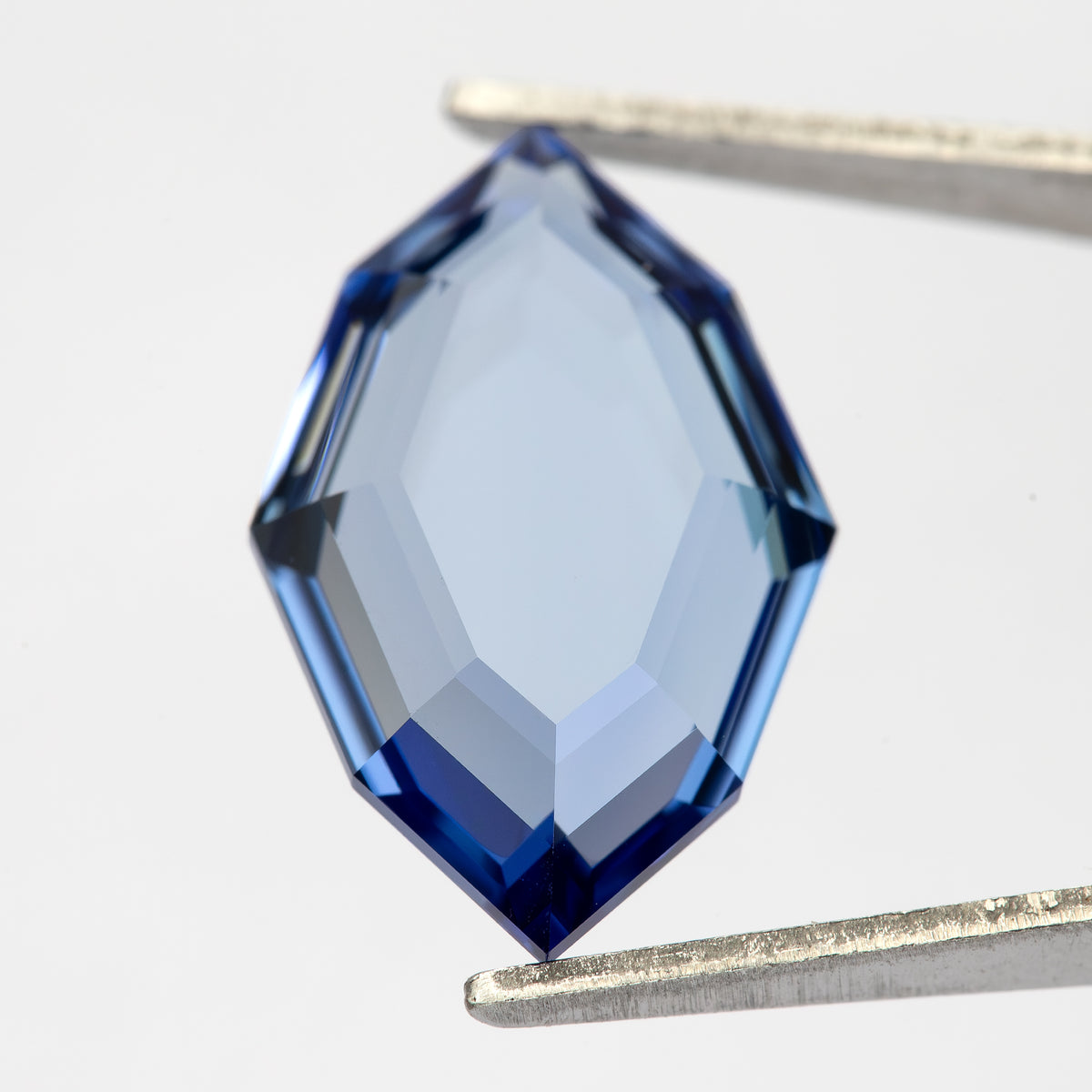 Blue portrait-cut hexagonal sapphire held face-up in tweezers against white, with an open transparent centre.