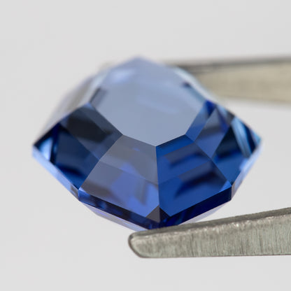 Angled pavilion-side view of the blue portrait-cut sapphire in tweezers, showing broad blue facets.
