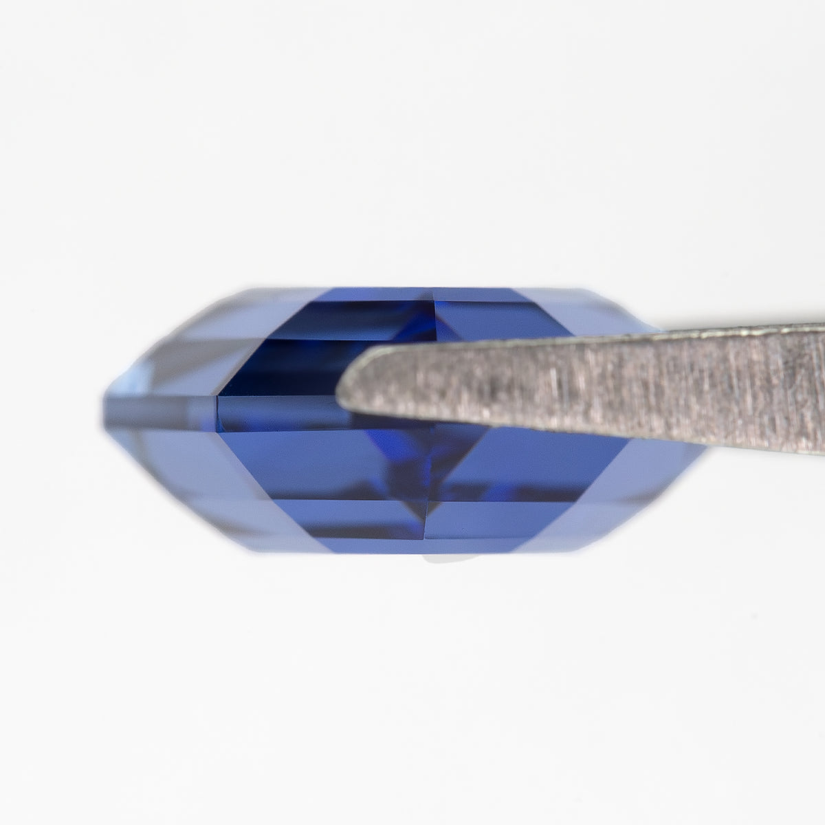 Side profile of the blue portrait-cut sapphire held horizontally in tweezers, showing its shallow depth.