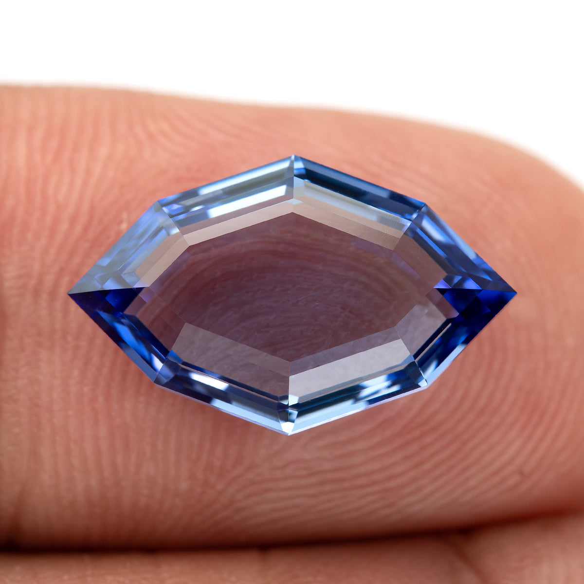 Blue portrait-cut sapphire resting face-up on fingertips, with the skin visible through the broad central table.