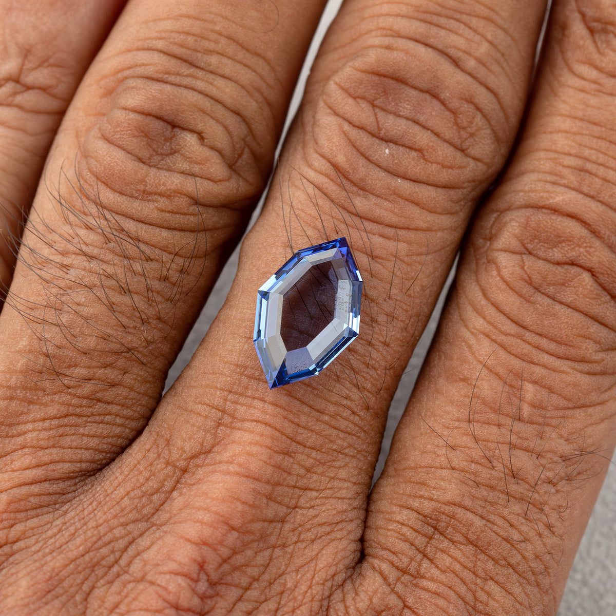 Blue portrait-cut sapphire resting on the back of a hand, showing its face-up size relative to the fingers.