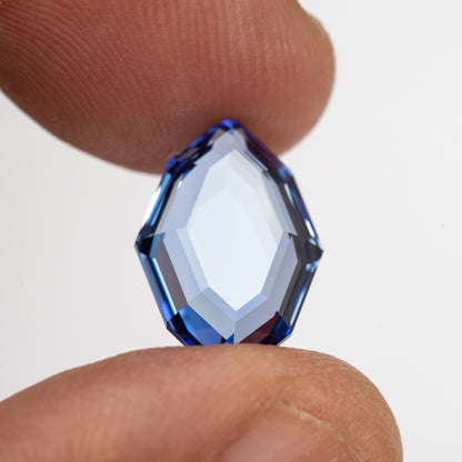 Blue portrait-cut sapphire held upright face-up between fingertips for scale.