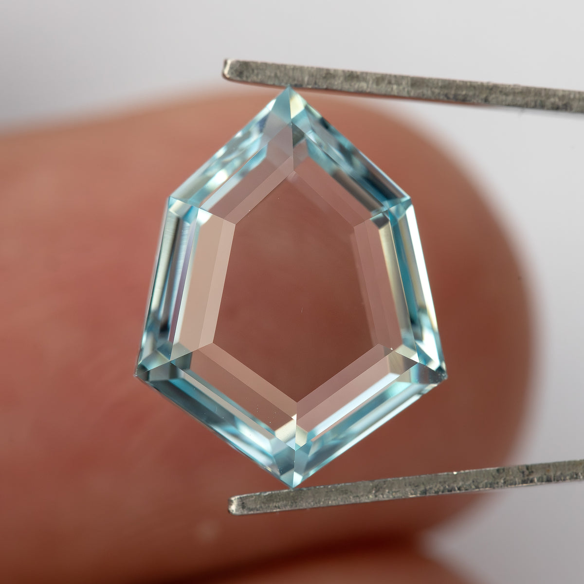 Aquamarine-blue shield-shaped lab-grown sapphire held in tweezers against a finger. View 6.