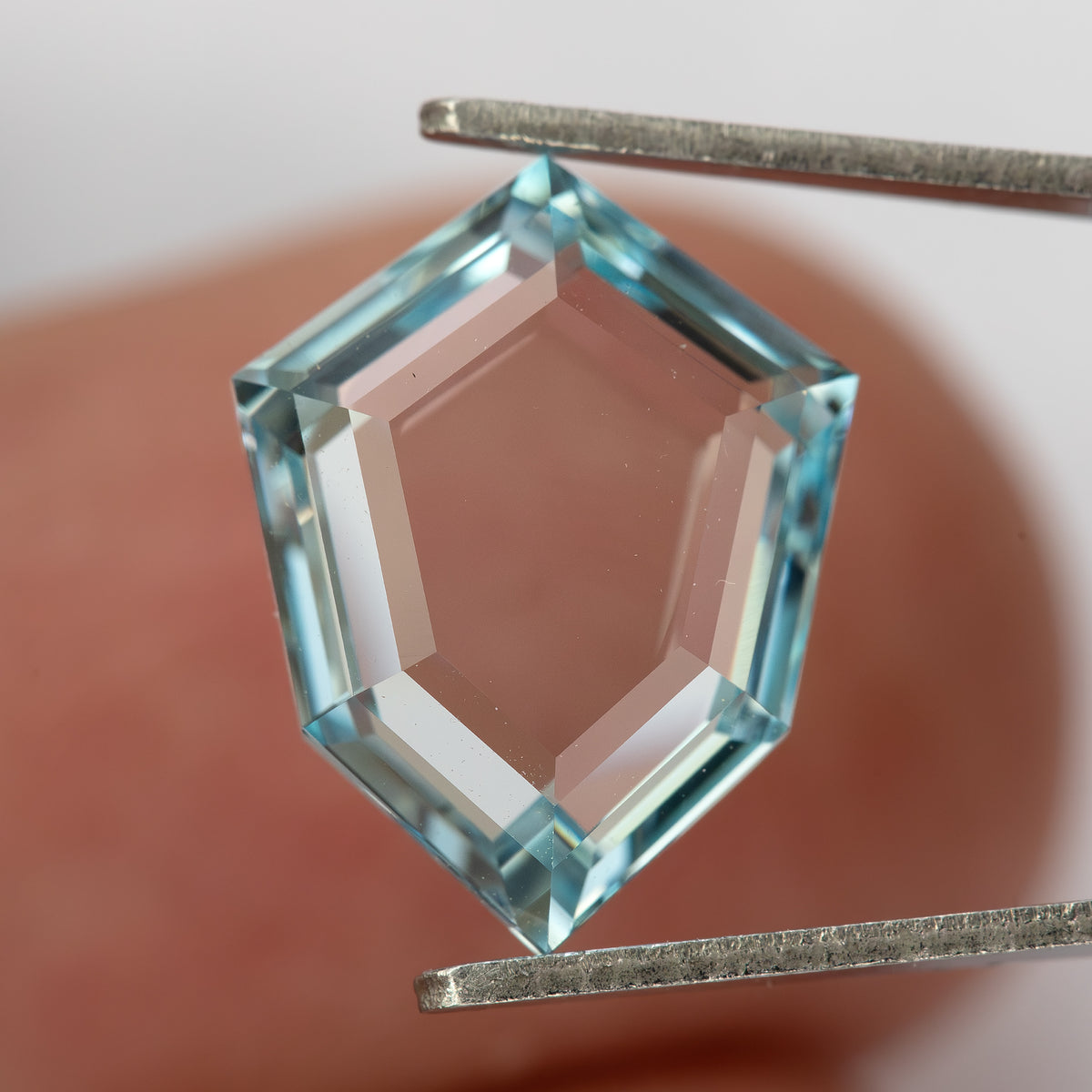 Aquamarine-blue shield-shaped lab-grown sapphire held in tweezers against a finger. View 7.