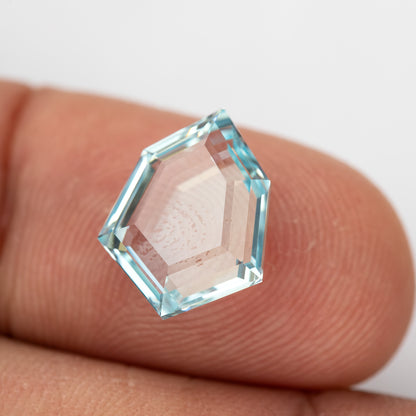 Aquamarine-blue shield-shaped lab-grown sapphire resting on fingertips for scale. View 15.