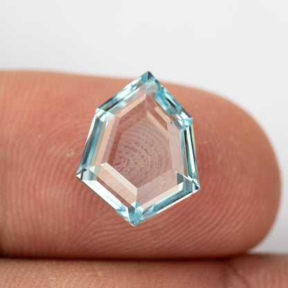 Aquamarine-blue shield-shaped lab-grown sapphire resting on fingertips for scale. View 16.