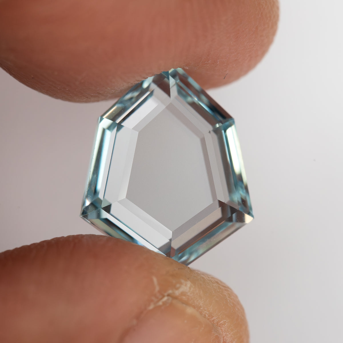 Aquamarine-blue shield-shaped lab-grown sapphire held between fingertips for scale. View 3.