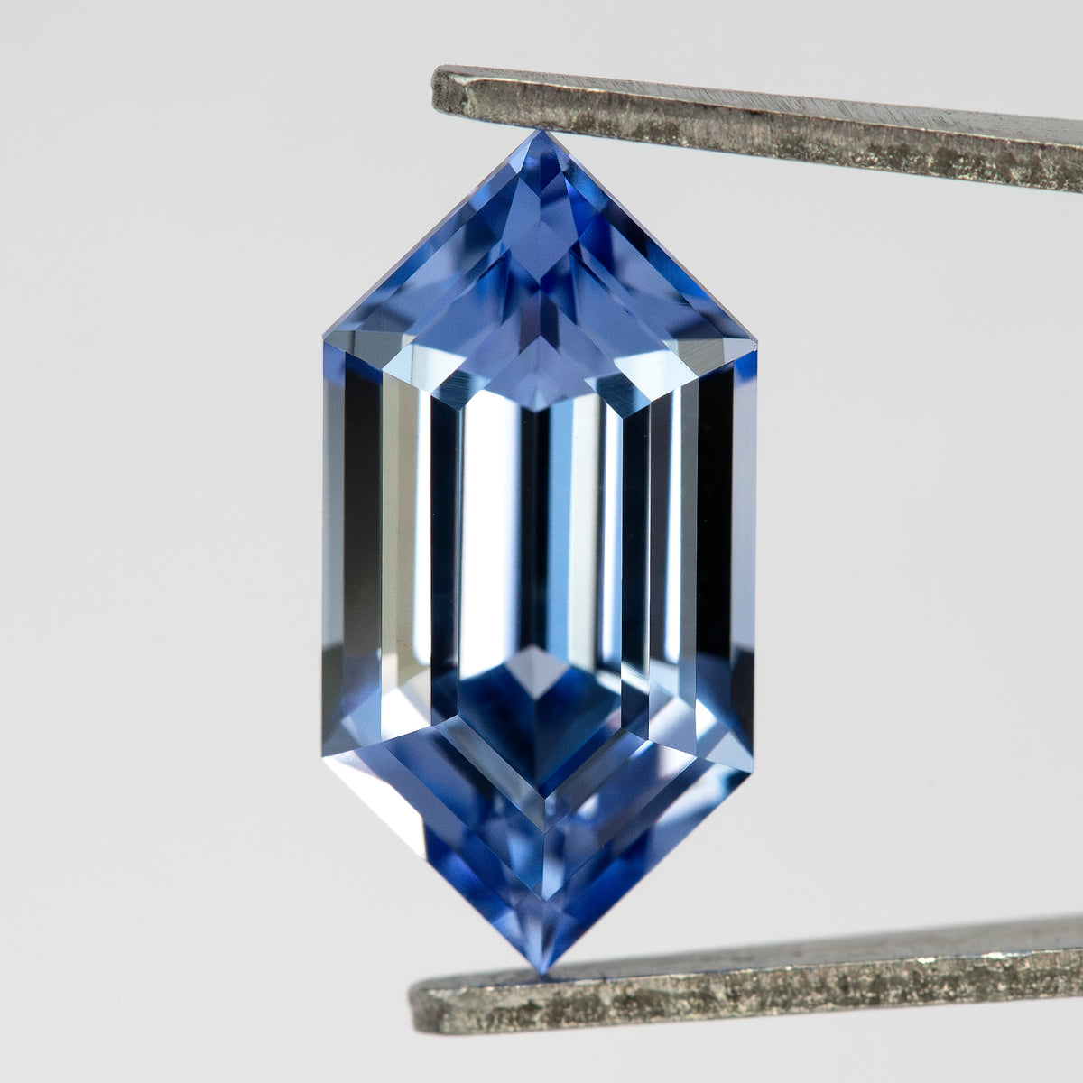 Blue lab-grown sapphire held upright in tweezers, showing the complete long-hexagon outline and step-cut face.