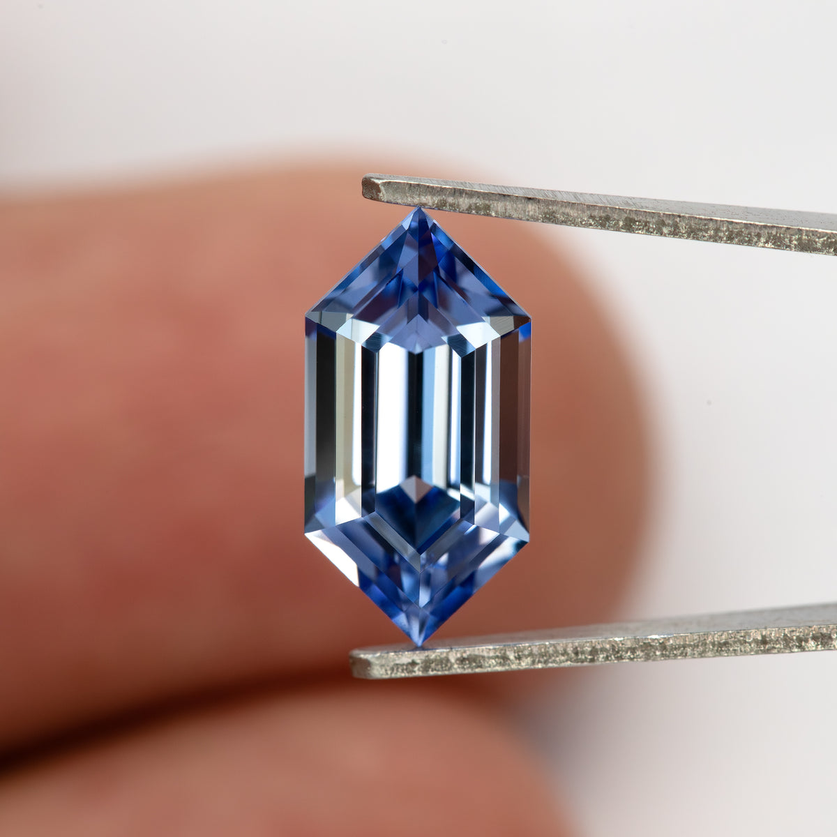 The blue long-hexagon sapphire face-up in tweezers, with a fingertip behind it for scale.