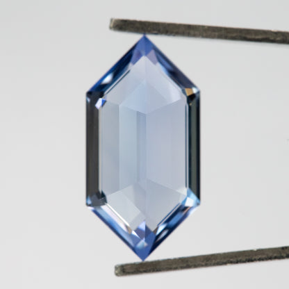 Reverse of the blue elongated-hexagon sapphire held upright in tweezers against a pale background.