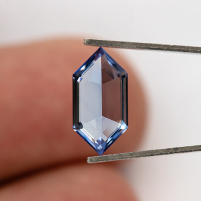 The underside of the blue long-hexagon sapphire in tweezers, with a fingertip behind it for scale.