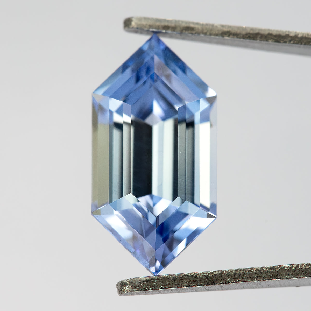 Blue step-cut sapphire in tweezers at a slight angle, showing pale central reflections and blue pointed ends.