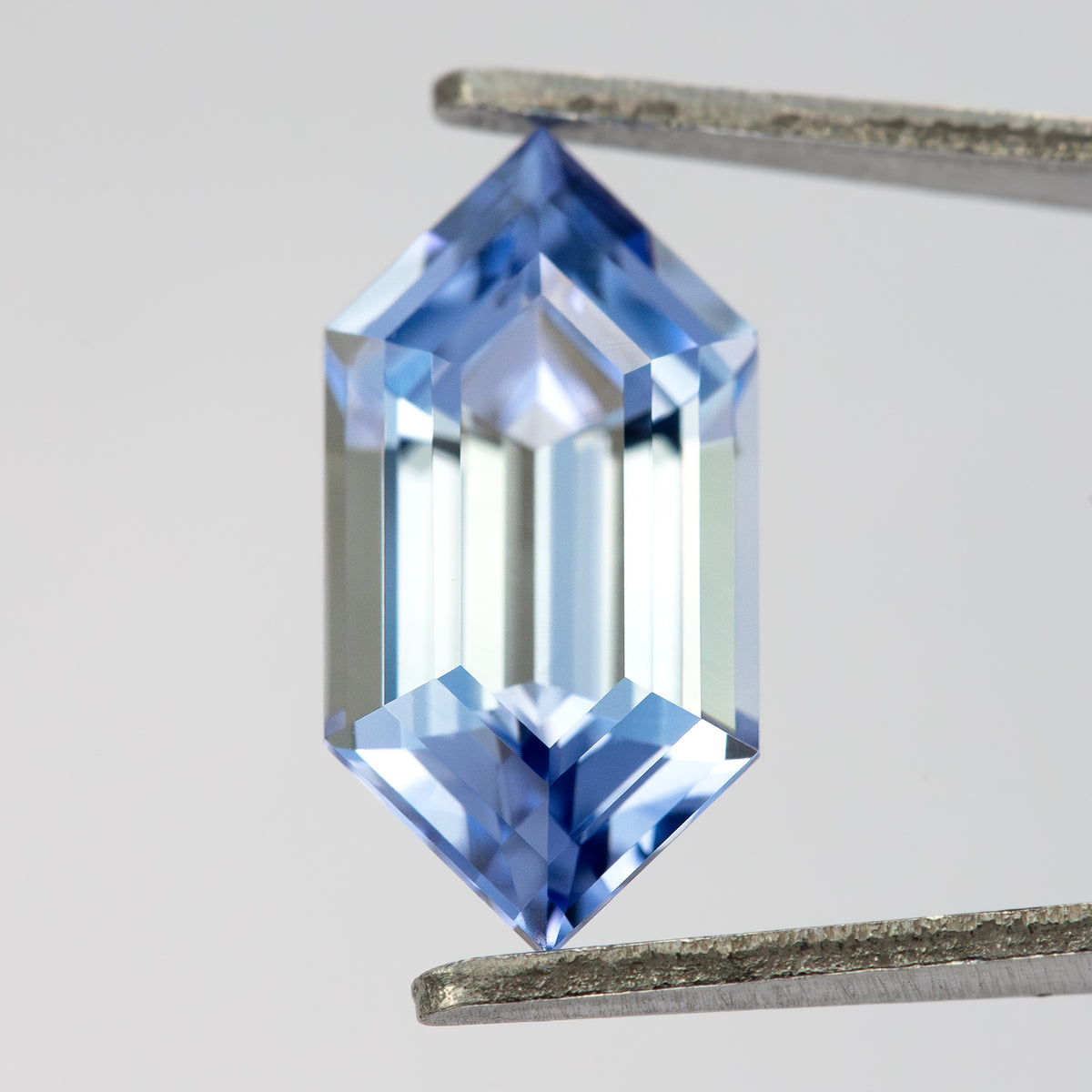 Upright blue long-hexagon sapphire in tweezers, with bright long reflections running through the center.